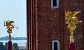 Two of three sword wielding golden lion heralds atop red flag posts in Piazza San Marco.
